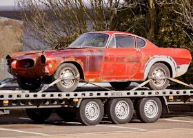 A worn-out, rusty vintage car secured on a flatbed trailer, outdoors under daylight.